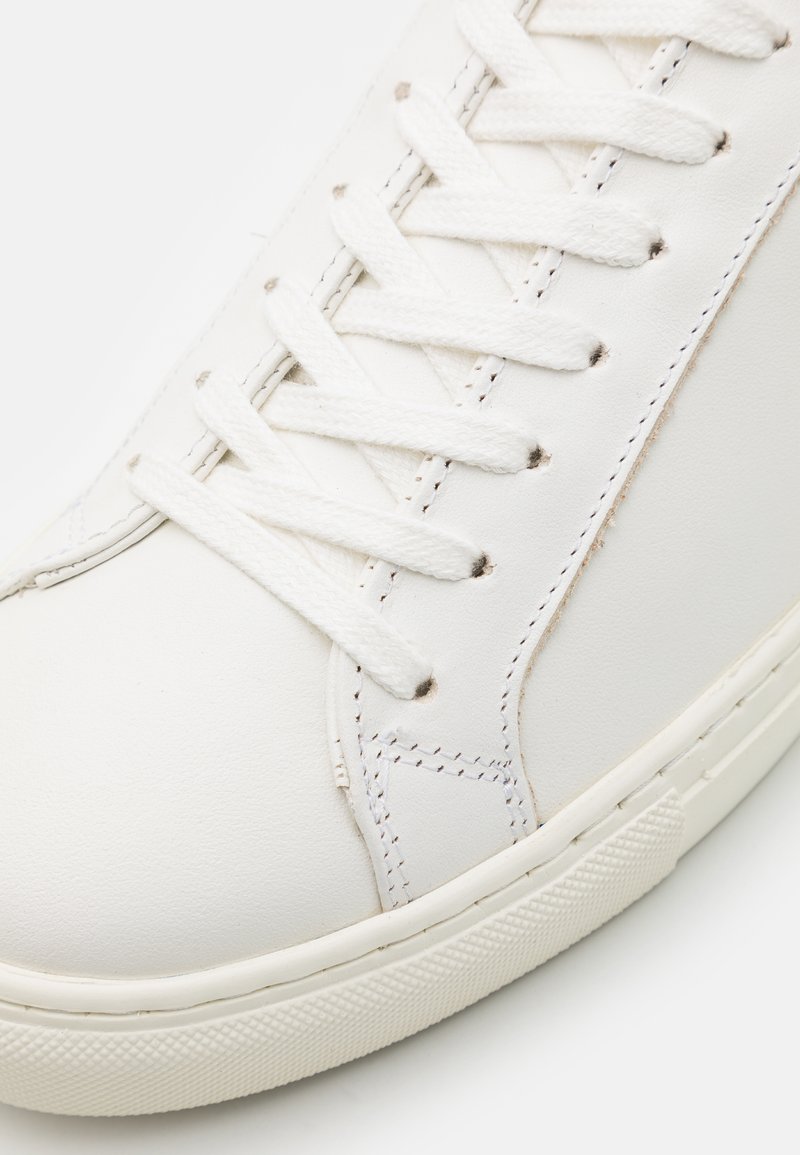 Baskets blanches en cuir avec une texture lisse, un design classique à lacets et des détails de couture. Semelle en caoutchouc avec un motif de grip texturé.