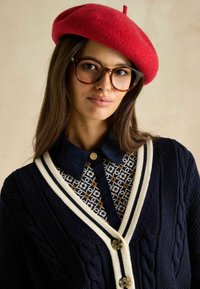 Jūras spēku adīts džemperis ar kontrastējošu balto apmalīti un zelta pogām, uzvilkts virs rakstainas krekla ar tumšu apkakli, apvienots ar sarkanu bereti.