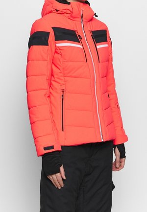 Pikowana kurtka neonowa pomarańczowa z czarno-białymi paskami na piersi, z zamkiem błyskawicznym z przodu, kieszeniami na zamek oraz mankietami z otworami na kciuki, noszona z czarnymi spodniami.