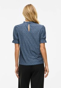 Blauwe geborduurde blouse met poffende korte mouwen, keyhole rugdetail, en kantachtige patronen. Gladde textuur; losse pasvorm.