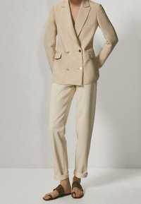 Beige Doppelreiher-Blazer mit goldenen Knöpfen, kombiniert mit cremefarbenen, taillierten Hosen und braunen geflochtenen Sandalen. Glatte Textur, maßgeschneiderte Passform.
