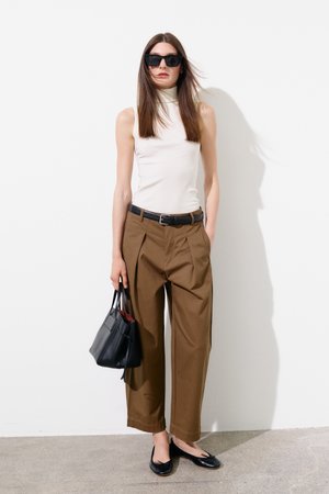 Femme aux longs cheveux bruns portant des lunettes de soleil noires, un col roulé blanc sans manches, un pantalon plissé marron, une ceinture noire, des ballerines noires et tenant un sac à main noir.