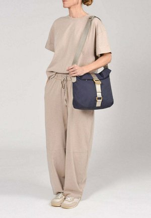 Persona que lleva una camiseta de talla suelta color beige y pantalones con zapatillas beige, llevando un bolso de hombro azul marino con correa beige y hebilla dorada.