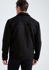 Giacca in suede nera con colletto, cucitura a yoke sul retro e dettagli con bottoni a pressione sui polsini. Texture morbida e vestibilità su misura.