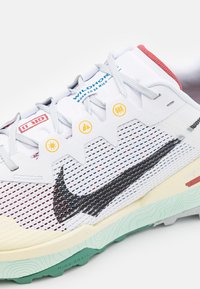 Zapatilla para trail running con parte superior de malla blanca, logo Swoosh negro, cordones grises y suela exterior verde menta con patrones texturizados para mayor agarre.