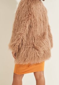 Mujer vestida con un abrigo de piel sintética marrón claro y desaliñado sobre una falda naranja, de espaldas con una mano visible a su lado.
