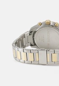 Montre en acier inoxydable argenté et doré avec un bracelet en métal, affichant un cadran chronographe et un dos lisse. Les éléments notables incluent des boutons dorés.
