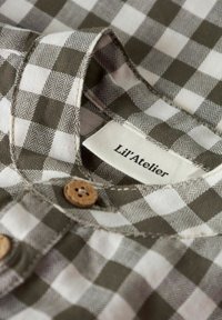 Žalias ir baltas languotas medvilninis audinys su žuvies kaulų raštu, su mediniu mygtuku ir etikete „Lil’Atelier“ šalia apykaklės.