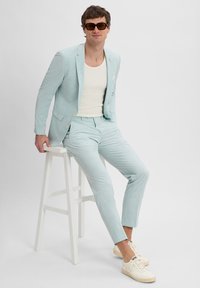 Helles, blaues Leinenanzug mit strukturiertem Finish, kombiniert mit einem weißen Tanktop und cremefarbenen Sneakern. Modell trägt dunkle Sonnenbrille.