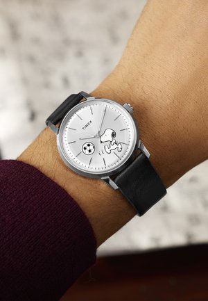 Ura z ročno uro s črnim usnjenim trakom, belim številčnico, na kateri je Snoopy, kako brca nogometno žogo, v bordo rdečem rokavu, čas 10:07:05.