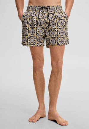 Man draagt een korte broek met zwarte, gele en witte geometrische patronen, handen in de zakken, staat op blote voeten op een grijze vloer.