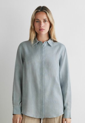 Kvinde med blondt hår iført en lyseblå skjorte med lange ærmer og knapper, stående mod en ensfarvet hvid baggrund.