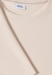 Beige kortærmet skjorte med rund halsudskæring, foldede ærmer og hvidt label med mærkenavnet "CECIL."