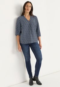Marinebluse mit geometrischem weißem Muster, V-Ausschnitt, 3/4-Ärmel. Dunkle Jeans im Skinny-Stil. Schwarze Ankle Boots. Schlichte, klare Ästhetik.