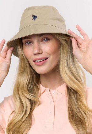 Beežist värvi kaanega müts mereväe sinise logoga. Mütsil on lai ääreosa ja sile pind, mis on kombineeritud heleroosa kraega särgiga.