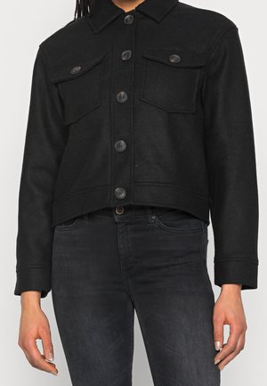 Veste noire courte à boutons avec poches poitrine à rabat portée sur un jean noir, visible du cou jusqu'aux hanches sur une personne.