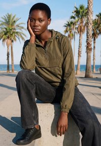 Mladić u maslinastoj jakni i crnim trapericama sjedi na betonskom bloku, oslanjajući glavu na ruku, s palmama i oceanom u pozadini.