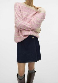 Personne portant un pull oversize rose et blanc à motifs, une jupe en denim foncé, des chaussettes grises et des bottes marron montantes sur un fond blanc.