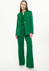 Blazer verde su misura e pantaloni wide-leg abbinati, realizzati in tessuto morbido, con un design a chiave sul collo e hardware dorato sottile.