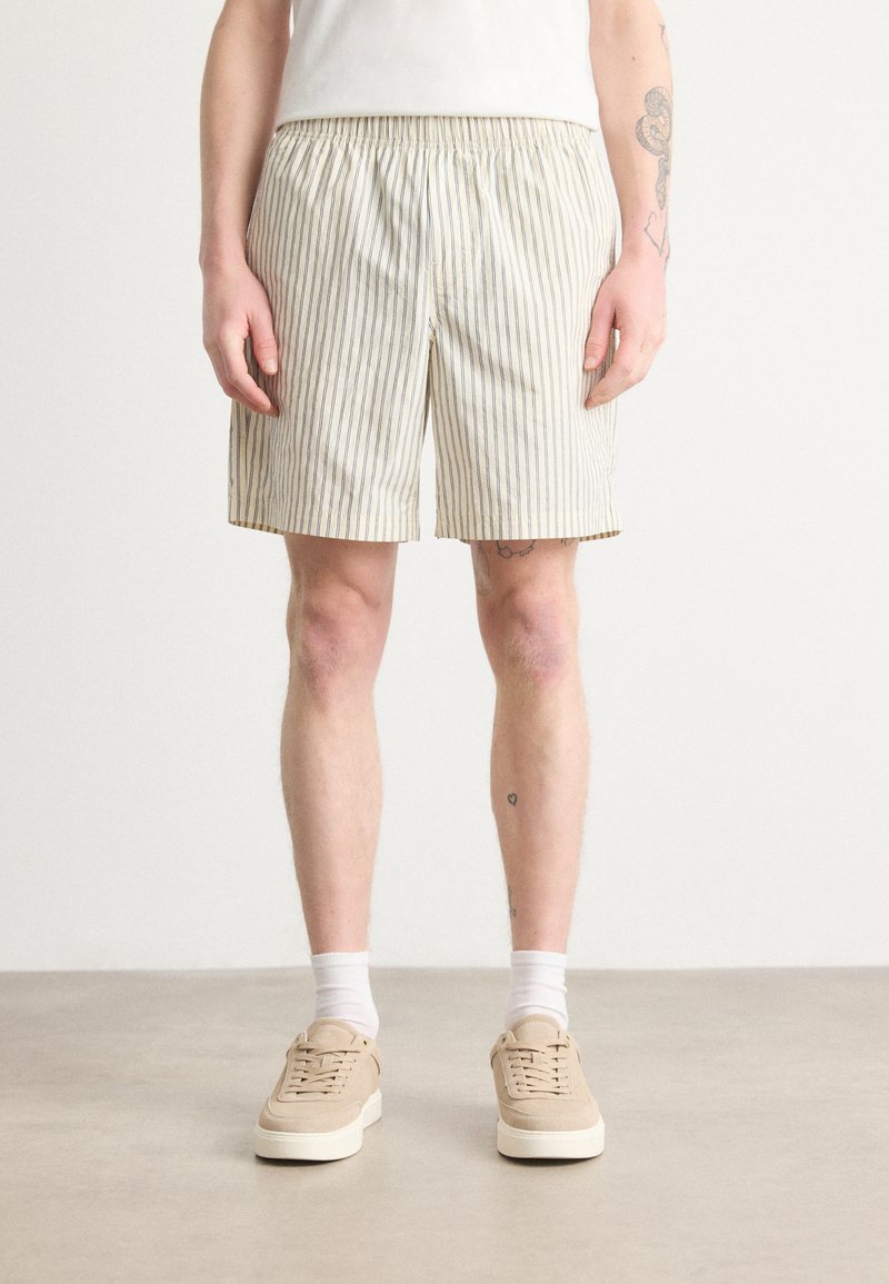 Pantalones cortos rayados beige con cintura elástica, frente plano y corte a la altura de la rodilla, combinados con zapatillas beige claro y calcetines blancos.