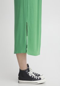 Rochie midi verde cu pliuri verticale și slit pe lateral, asortată cu teniși negri cu guler înalt, având talpa și șireturile albe.