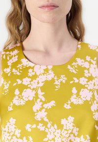 Bluză fără mâneci, cu guler rotund, de culoare galben-muștar, cu un model floral roz. Materialul are o textură ușoară și un finisaj neted.