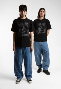 Sorte bomull T-skjorter har et grafisk design i grått. Kombinert med løse blå denimbukser og svarte joggesko. Flatt, litt oversized passform.
