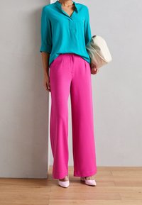 Türkisfarbene Bluse mit V-Ausschnitt und aufgerollten Ärmeln, kombiniert mit weit geschnittenen rosa Hosen. Das Model hält eine cremefarbene Tasche und trägt rosa Absätze.