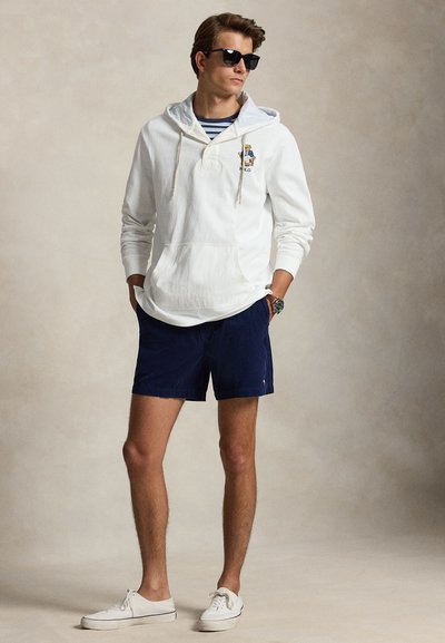 Homme portant des lunettes de soleil noires, un sweat à capuche blanc avec un logo d'ours, un short bleu marine, des baskets blanches et une chemise rayée en dessous, debout les mains dans les poches.