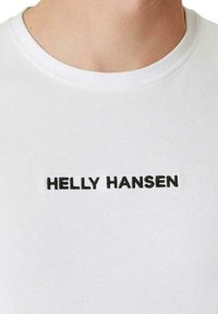 Camiseta de algodón blanca con cuello redondo y texto bordado en negro "HELly HANSEN" en el frente, mostrando una textura suave.