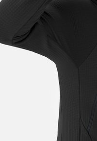 Gros plan sur la zone de l'aisselle et la couture latérale d'une veste noire texturée, avec une fermeture éclair verticale visible sur un fond uni.