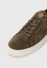 Baskets en daim marron avec un bout arrondi, une semelle en caoutchouc et des lacets plats. Texture remarquable sur la partie supérieure et branding subtil sur le côté.