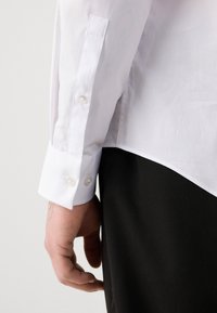 Weißes Langarmhemd mit geknöpften Manschetten und einer glatten Textur. Sichtbare Hand hält das Hemd in der Nähe des Saums. Schwarzer Stoff darunter.