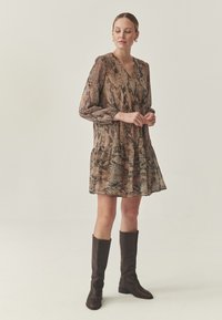 Braunes und beiges Kleid mit Schlangenmuster, langen Ärmeln, V-Ausschnitt und gerüschtem Saum, kombiniert mit knielangen braunen Lederstiefeln.