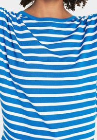 Modrý a bílý pruhovaný top s měkkou texturou, s širokým výstřihem a volným střihem, zdůrazňující horizontální linie.