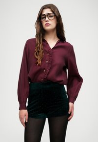 Βουργουνδί μπλούζα με κουμπιά και λεπτομέρειες βολάν, με μακριά μανίκια, συνδυασμένη με σκούρα πράσινα βελούδινα σορτς με μικρές βούλες. Φορεμένες μαύρες καλσόν.