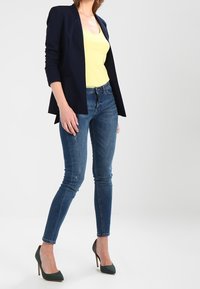 Navy blazer over a yellow tank top, paired with blue skinny jeans and green high heels. The blazer features structured shoulders and a smooth texture.