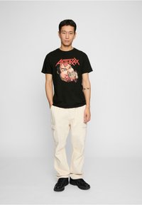 Sort bomulds T-shirt med et farverigt grafisk print med ordet "ANTHRAX" og en dramatisk ansigtsillustration. Båret sammen med cremede cargobukser.