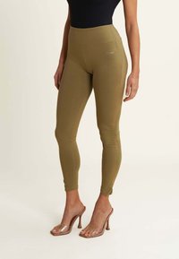 Leggings verde oliva con vita alta, realizzati in un materiale elasticizzato. Presentano un logo sul lato. Abbinati a sandali con tacco trasparente.