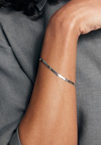 Metalowa bransoletka o zakrzywionym, teksturowanym wzorze. Polerowane wykończenie w kolorze srebrnym, noszona na ciemnoskórej ręce, na tle szarej tkaniny.