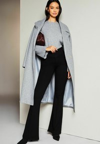 Manteau en laine gris porté sur un pull gris, associé à un pantalon évasé noir et un sac à main bordeaux, présentant une texture lisse et un design structuré.