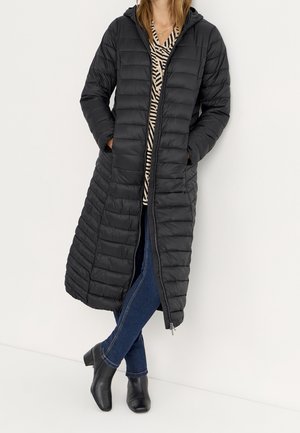 Vrouw draagt een lange zwarte doorgestikte capuchonjas, een blouse met patroon, blauwe spijkerbroek en zwarte enkellaarsjes, staand met handen in de zakken.