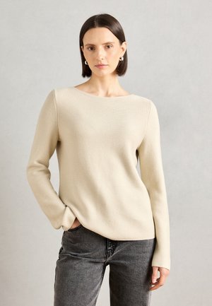 Lichtbeige trui met een bootnek, ribbeltextuur en lange mouwen, gedragen met grijze jeans, die een eenvoudig en modern ontwerp benadrukt.