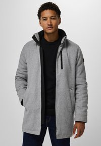 Homme portant un manteau zippé gris clair sur un pull noir et un jean foncé, se tenant contre un fond clair uniforme.