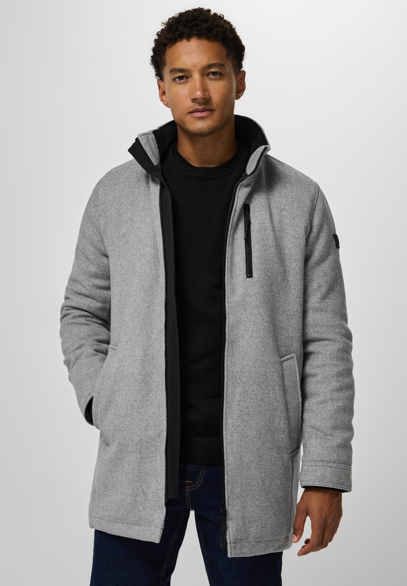 Homme portant un manteau zippé gris clair sur un pull noir et un jean foncé, se tenant contre un fond clair uniforme.