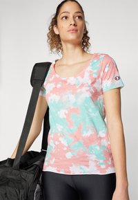 Camiseta de manga corta y escote en forma de barco con un patrón de tie-dye en rosa, verde y blanco. El tejido parece suave con un corte relajado. Logo en la manga.