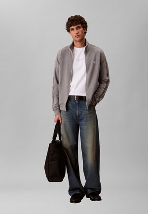 Mladý muž v sivej bunde na zips, bielom tričku, širokých modrých džínsoch a čiernych topánkach, držiaci čiernu tašku na rameno na jednoliatom sivom pozadí.