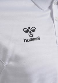 Polo blanc avec un logo abeille stylisé noir au-dessus du mot « hummel » sur la poitrine, tissu texturé et col à boutons visible.