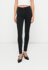 Jeans ajustados negros con una textura de denim suave, cintura de altura media y costuras en contraste a lo largo de las costuras laterales. Combinados con zapatos negros de punta.