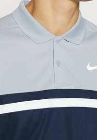 Šedé polo tričko s námořnickým pruhem a bílým akcentem. Vyrobeno z hladkého materiálu, má límeček a dva knoflíky. Bílé logo na hrudi.